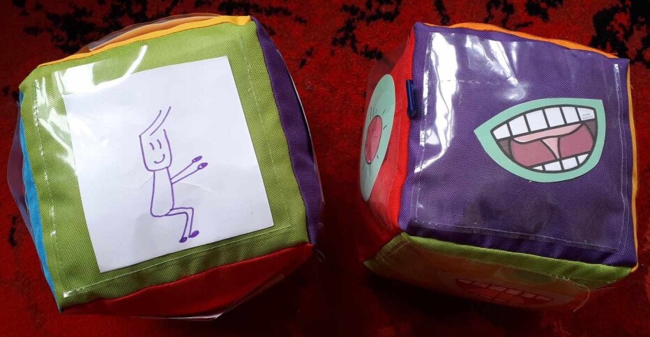 dwie kostki do gry z przezroczystymi kieszonkami z plastiku za którymi są rysunki ćwiczeń gimnastycznych - przysiady i ćwiczeń logopedycznych - kląskanie