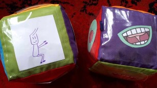 dwie kostki do gry z przezroczystymi kieszonkami z plastiku za którymi są rysunki ćwiczeń gimnastycznych - przysiady i ćwiczeń logopedycznych - kląskanie